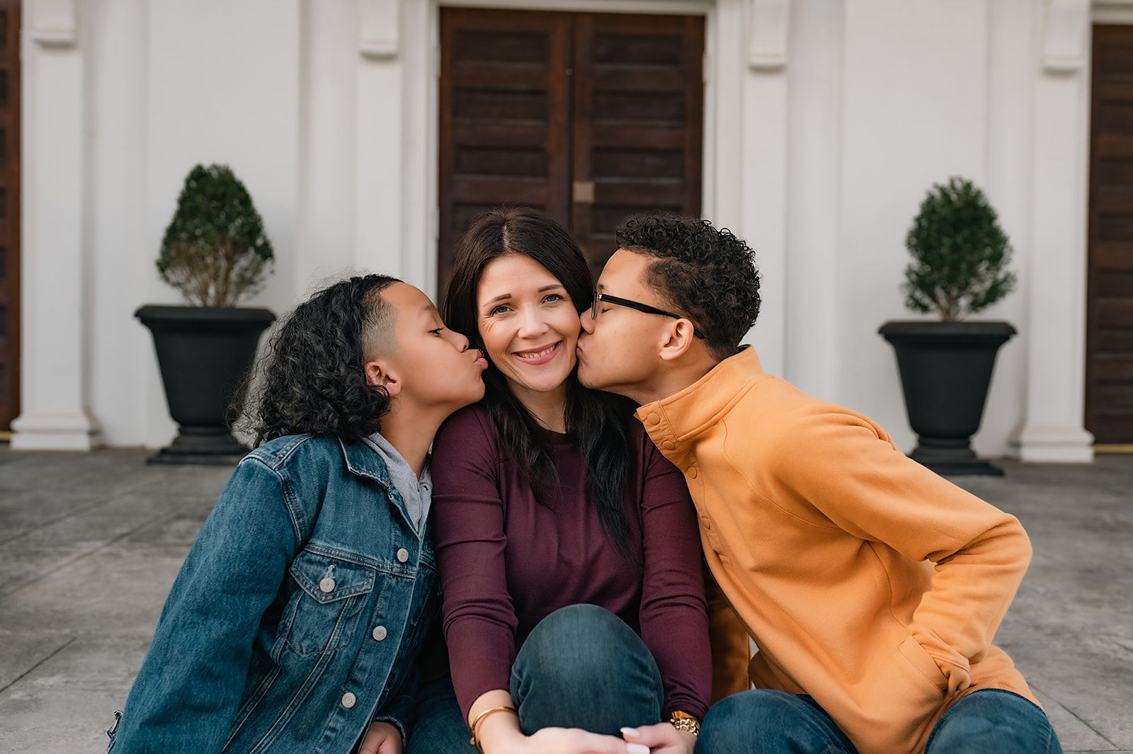 Three people sitting together share a sweet moment between green potted plants and a white building exterior.