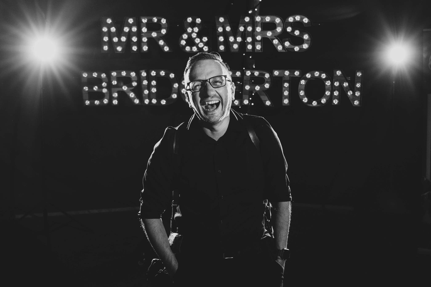 Man laughing in front of Mr & Mrs Bridgerton illuminated sign at wedding reception in black and white photo.