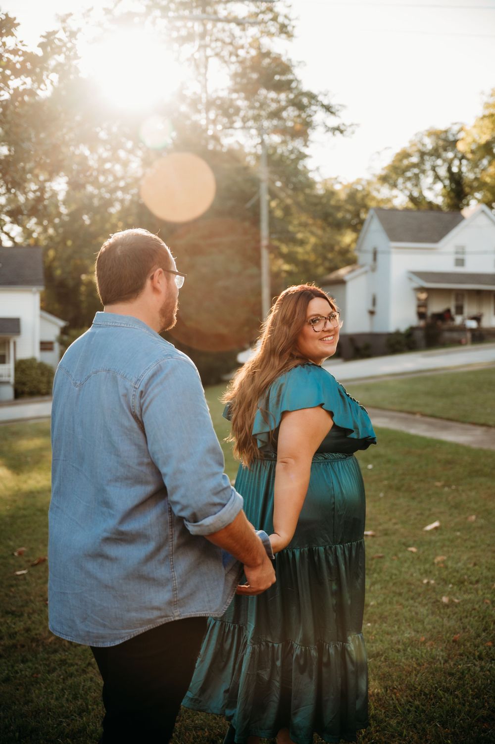 A couple stands together at sunset in a residential yard, with golden sunlight and lens flares in the background.