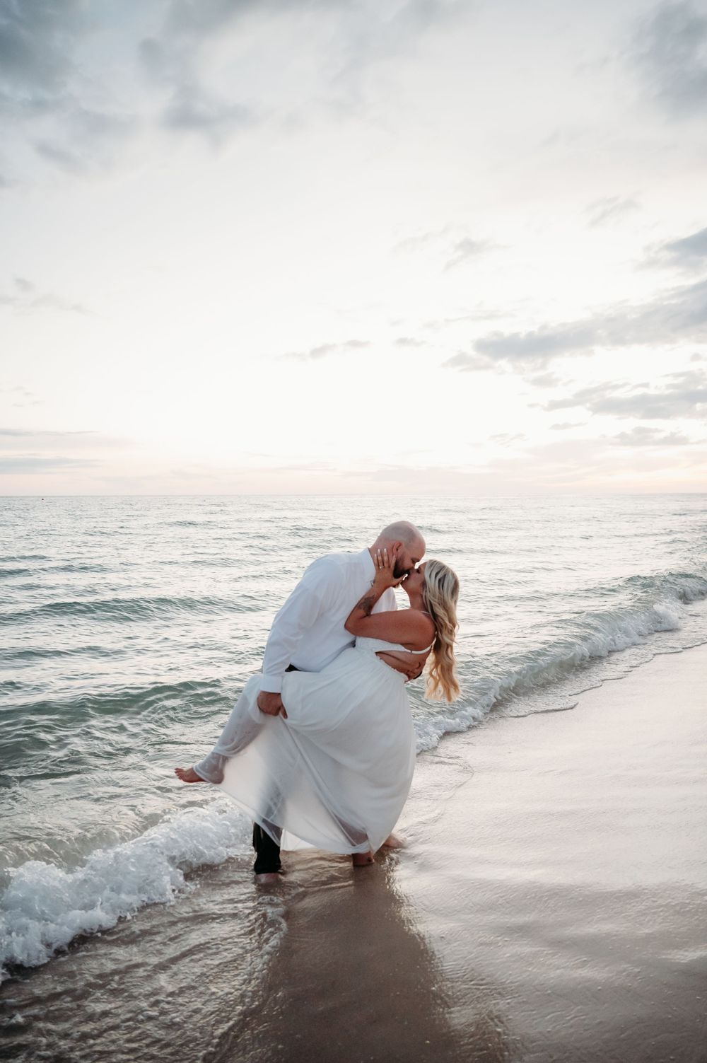 A romantic moment captured on a beach at sunset with waves lapping at the shore and soft cloudy skies above.