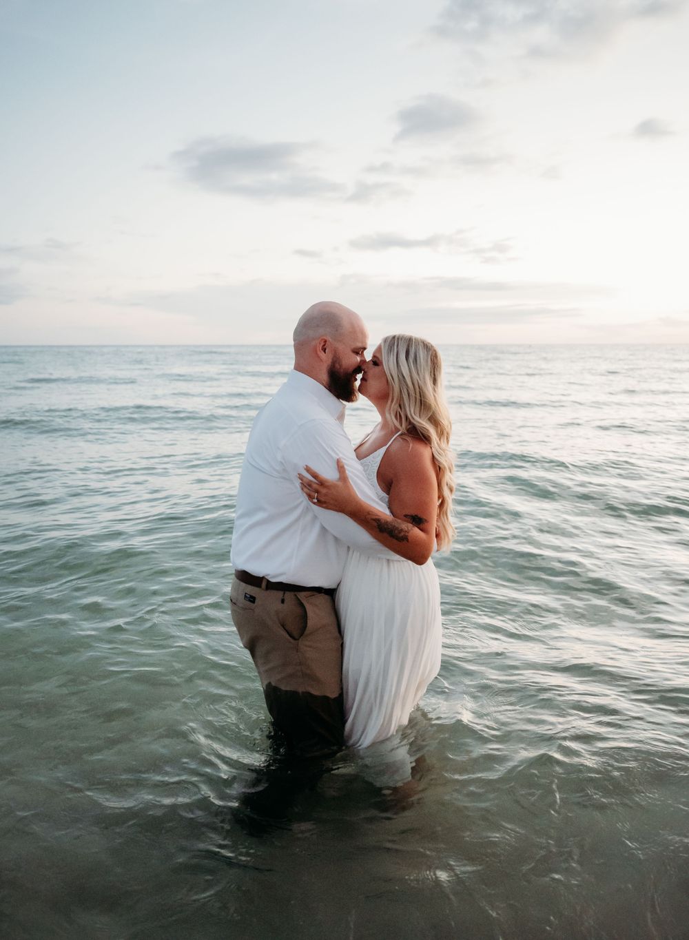 A romantic couple embraces while standing in the ocean water during sunset creating a dreamy beachside moment.