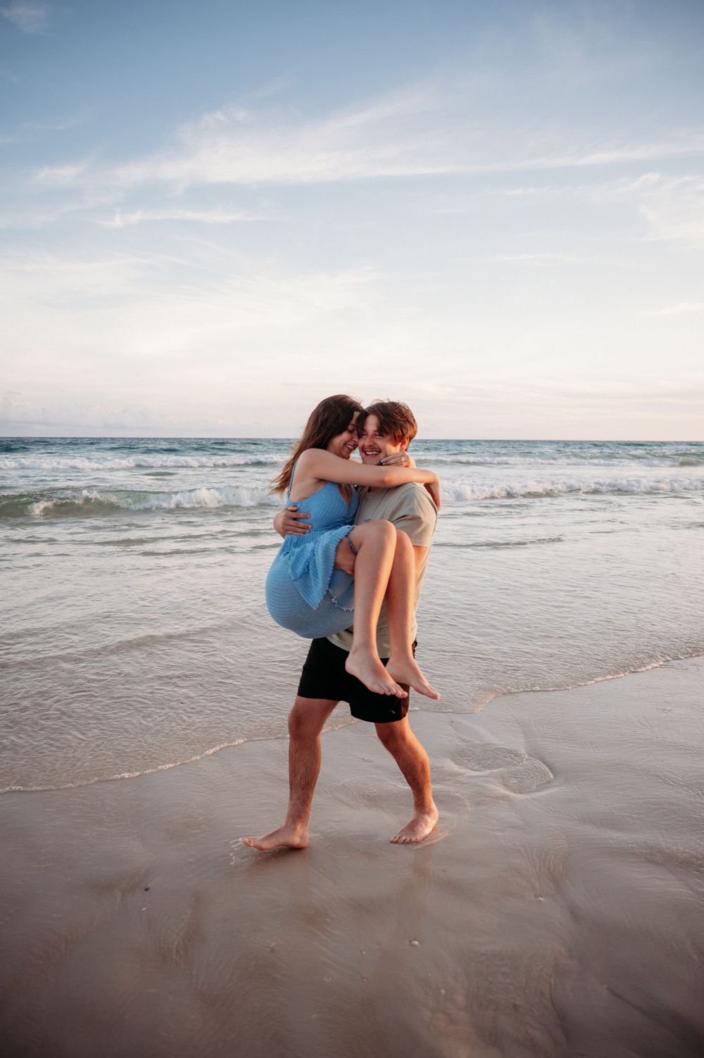 A couple embraces on a sandy beach at sunset as gentle waves roll onto the shore.
