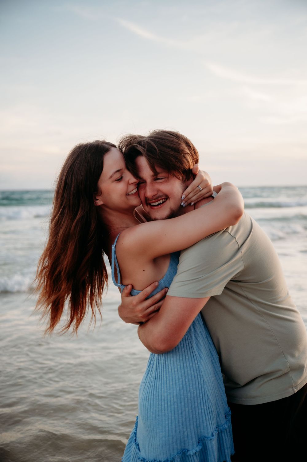 A romantic couple embraces and laughs together on the beach at sunset with ocean waves in the background.