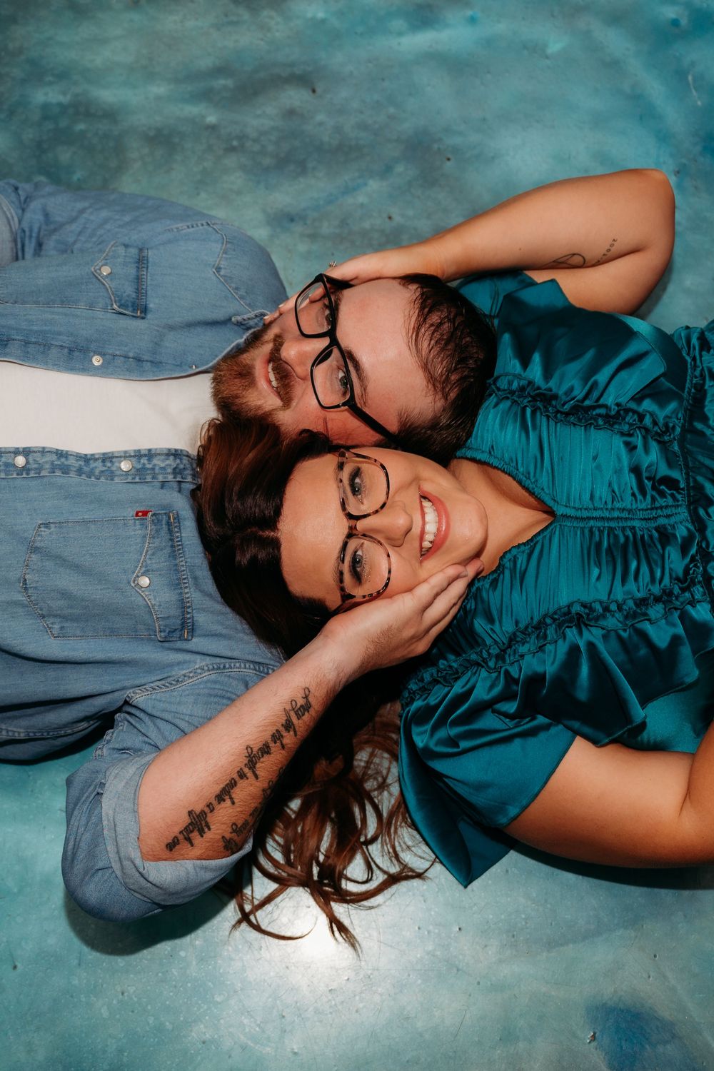 Two people wearing glasses lay together on a blue background, one in denim and one in a teal top.