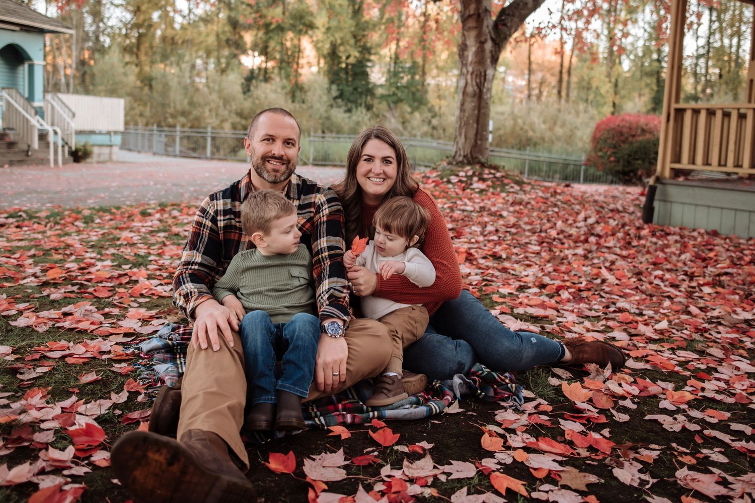 Family sitting together on red fallen autumn leaves in a backyard setting.