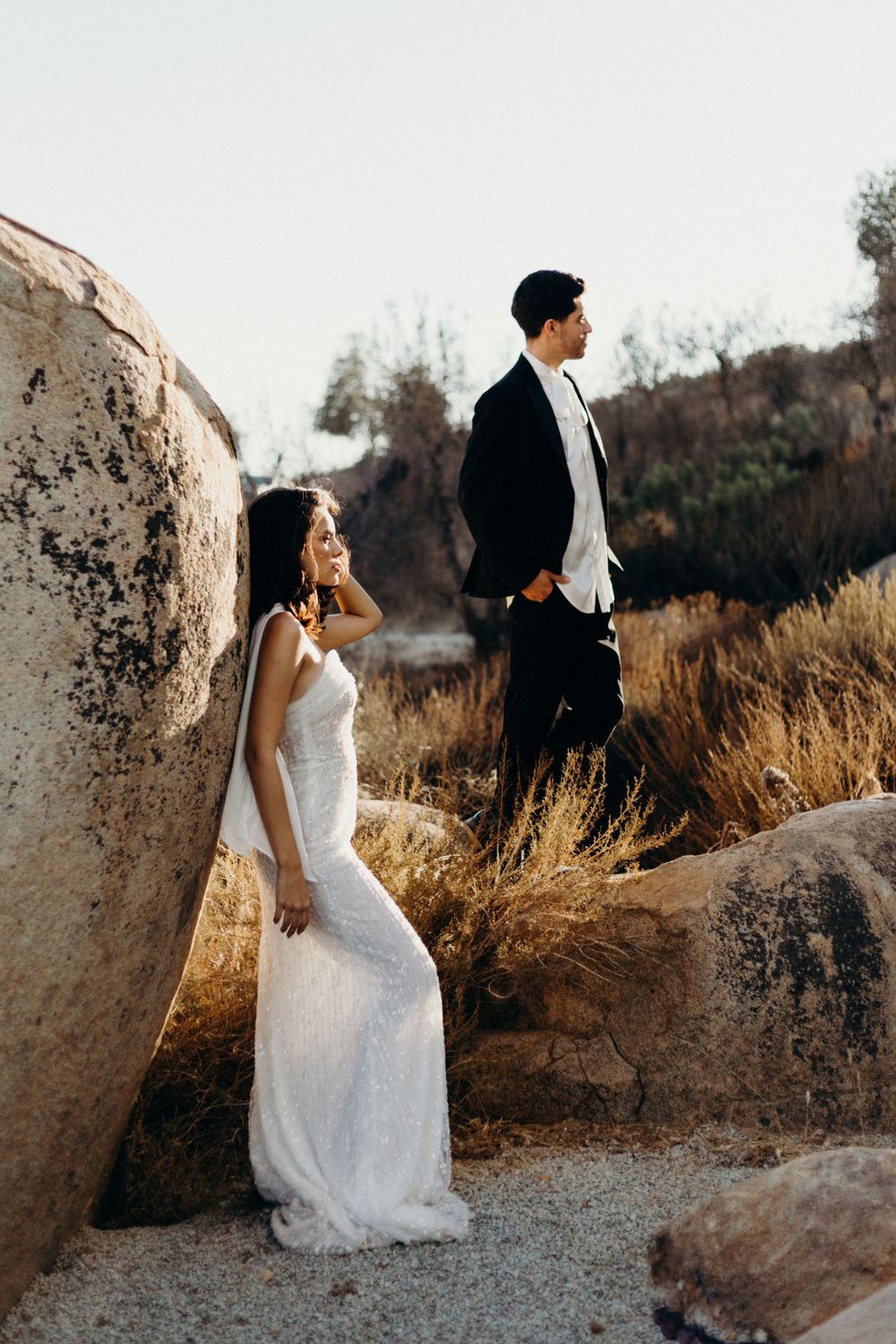 Couple in wedding attire pose among large boulders at sunset in a scenic natural setting.