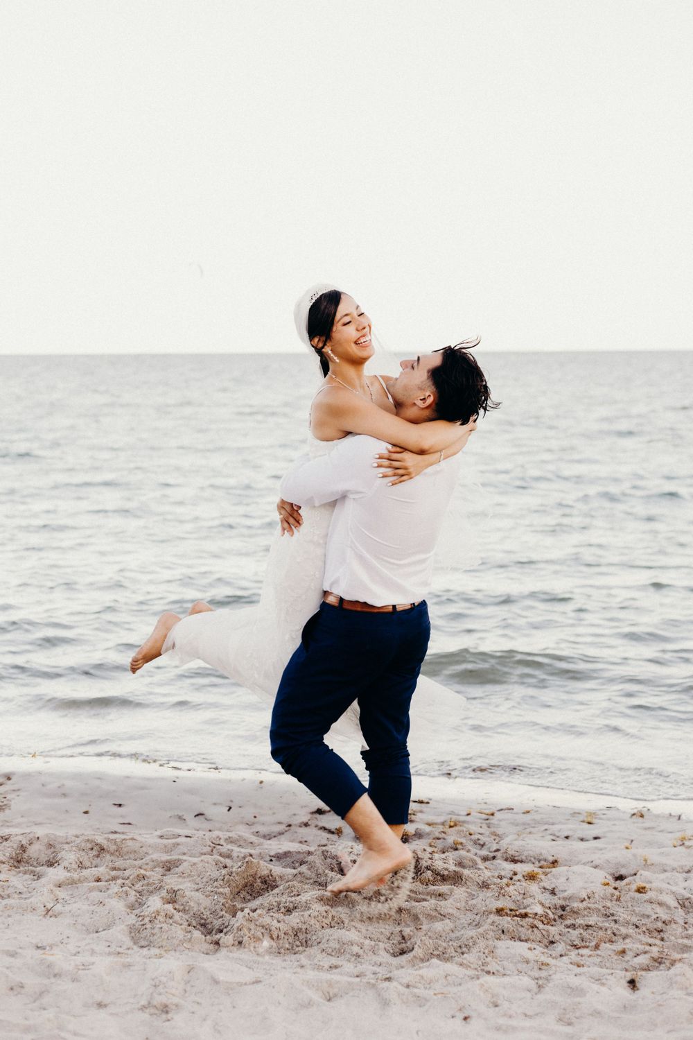 A romantic couple embraces joyfully on a sandy beach at sunset with ocean waves in the background.