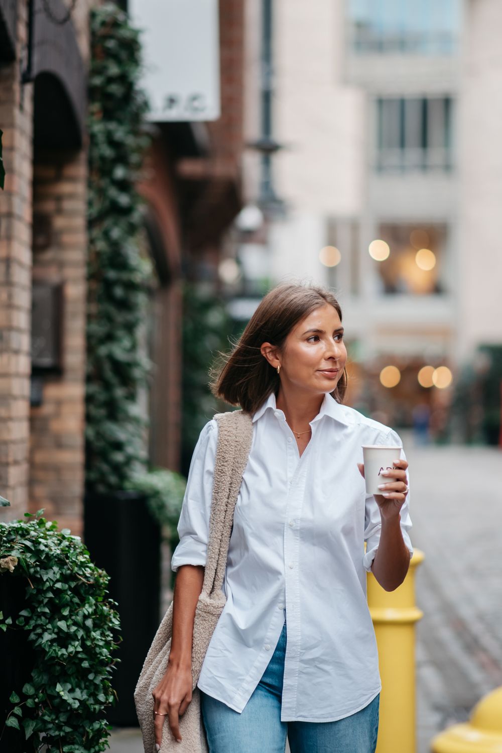 Woman in white button-down shirt and jeans walking on urban street with coffee cup and shoulder bag.