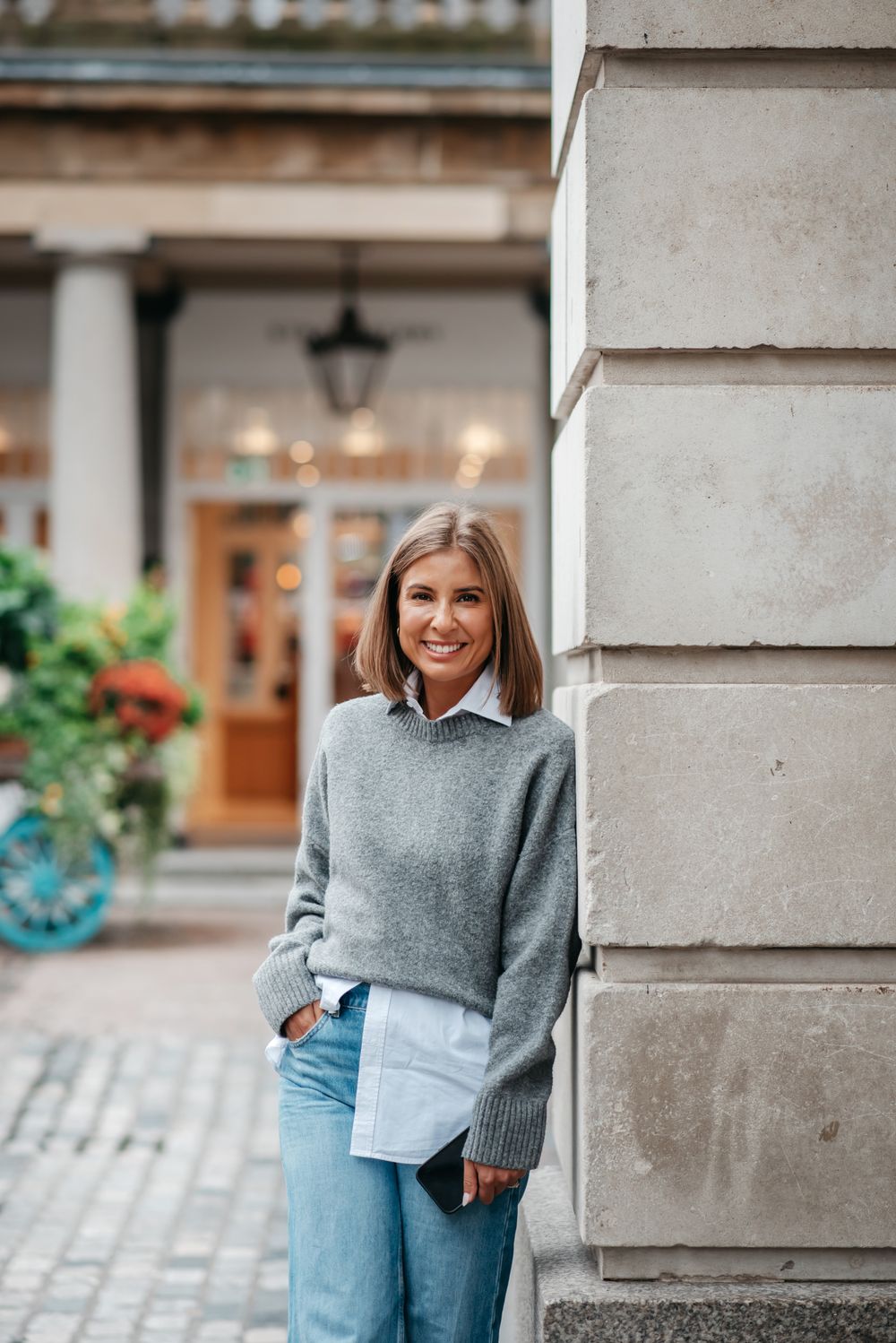 Person in casual streetwear outfit with grey sweater and jeans poses against stone building in urban setting.