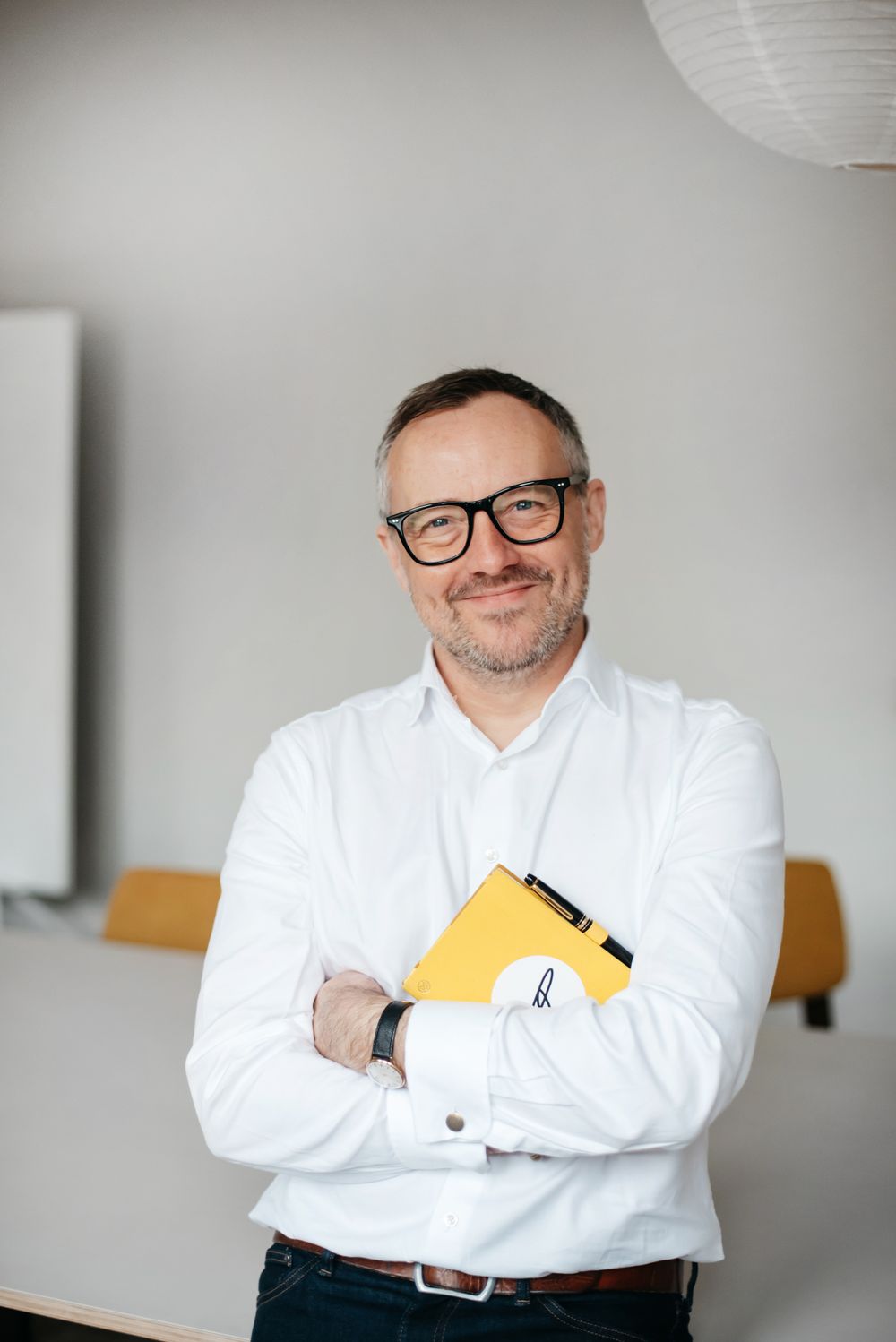 Professional in a white button-down shirt and glasses stands confidently holding a yellow folder in an office setting.