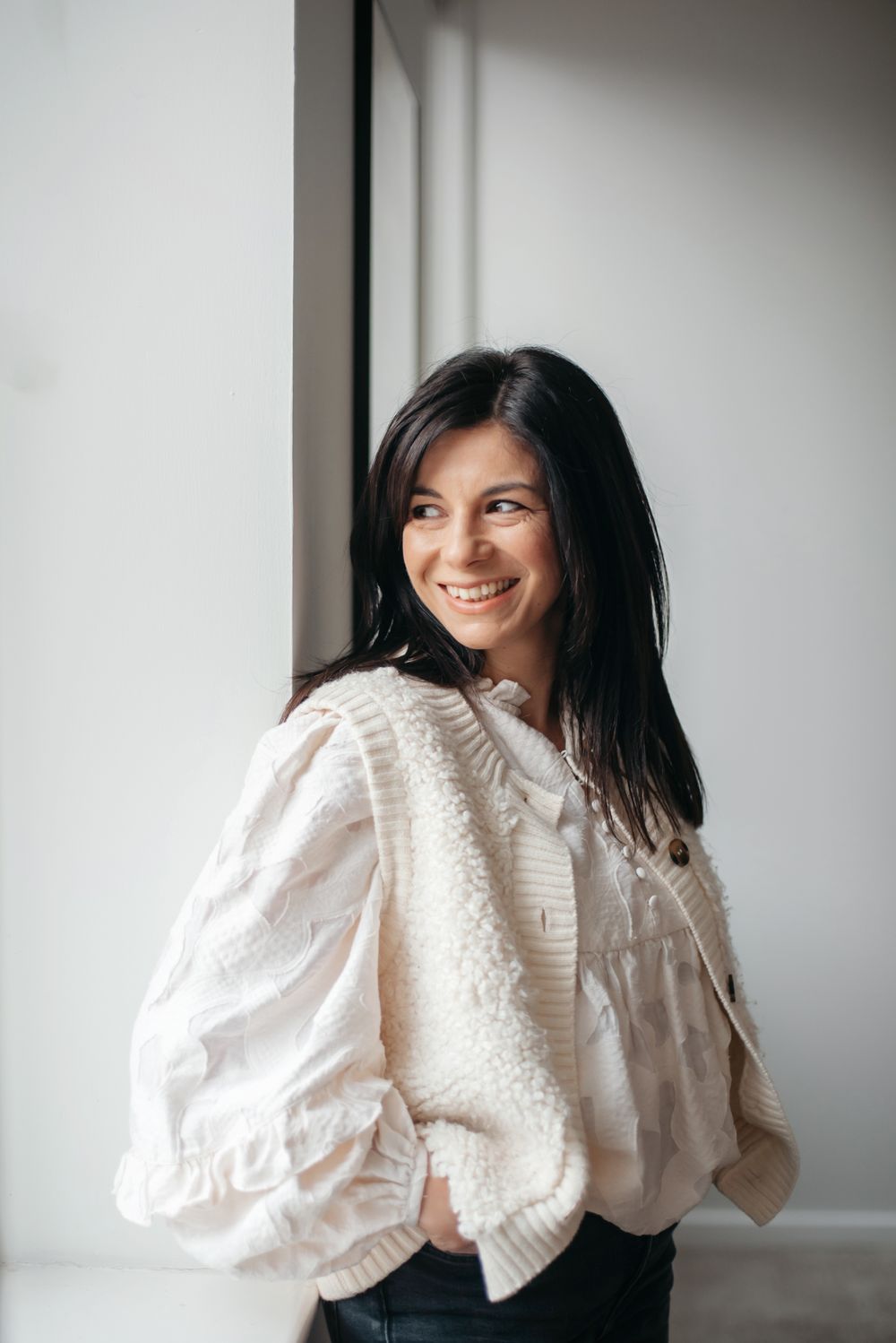 A person wearing a white knit sweater and dark pants smiles while standing near a window with natural lighting.