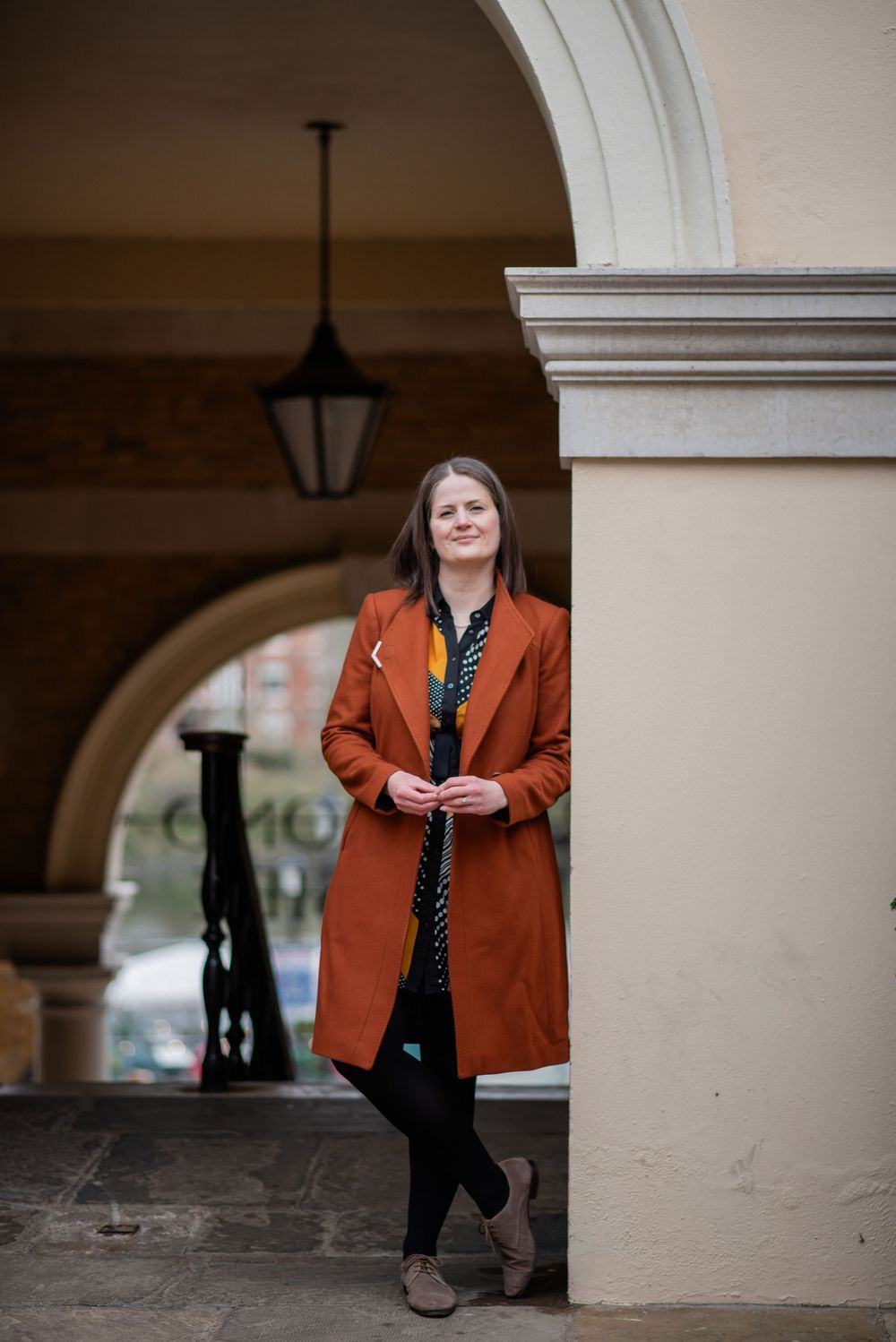 Someone wearing a burnt orange coat leans against a pillar beneath an arched passageway with a hanging lantern.