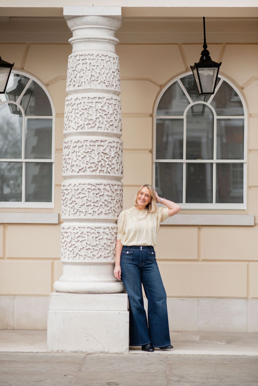 A person in flared jeans and a light colored top poses against an ornate classical column with arched windows in the background.