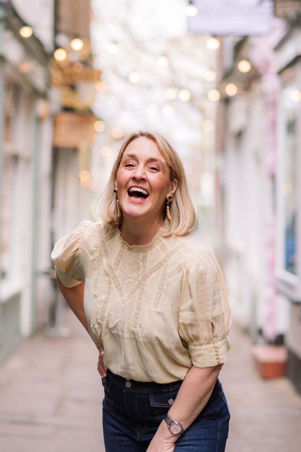 Person wearing cream colored blouse and jeans laughs joyfully in narrow alley with twinkling lights above.