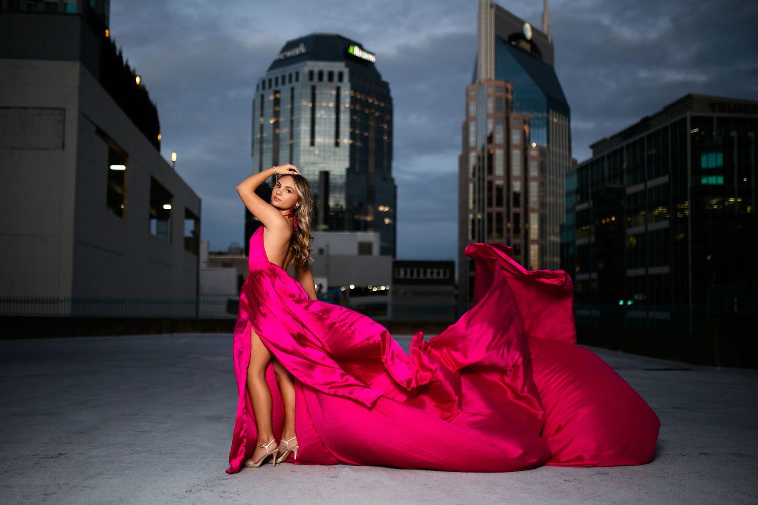 Senior girl in flowing pink dress poses dramatically against Nashville skyline at dusk. Middle TN senior photographer.