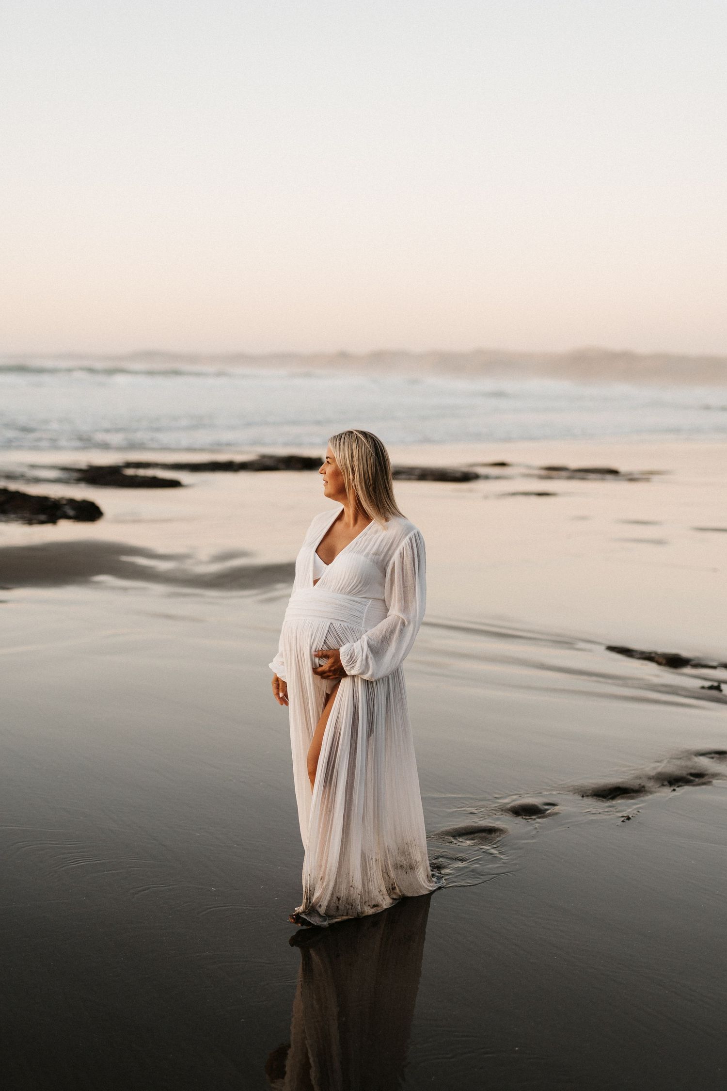 Ethereal maternity photo of an expectant mother in a flowing white dress standing on a misty beach at sunset.