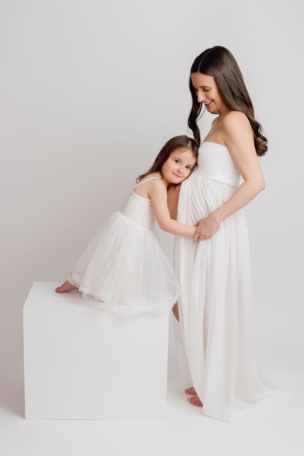Pregnant woman in white dress smiling at young girl in tutu hugging her belly in a bright studio.