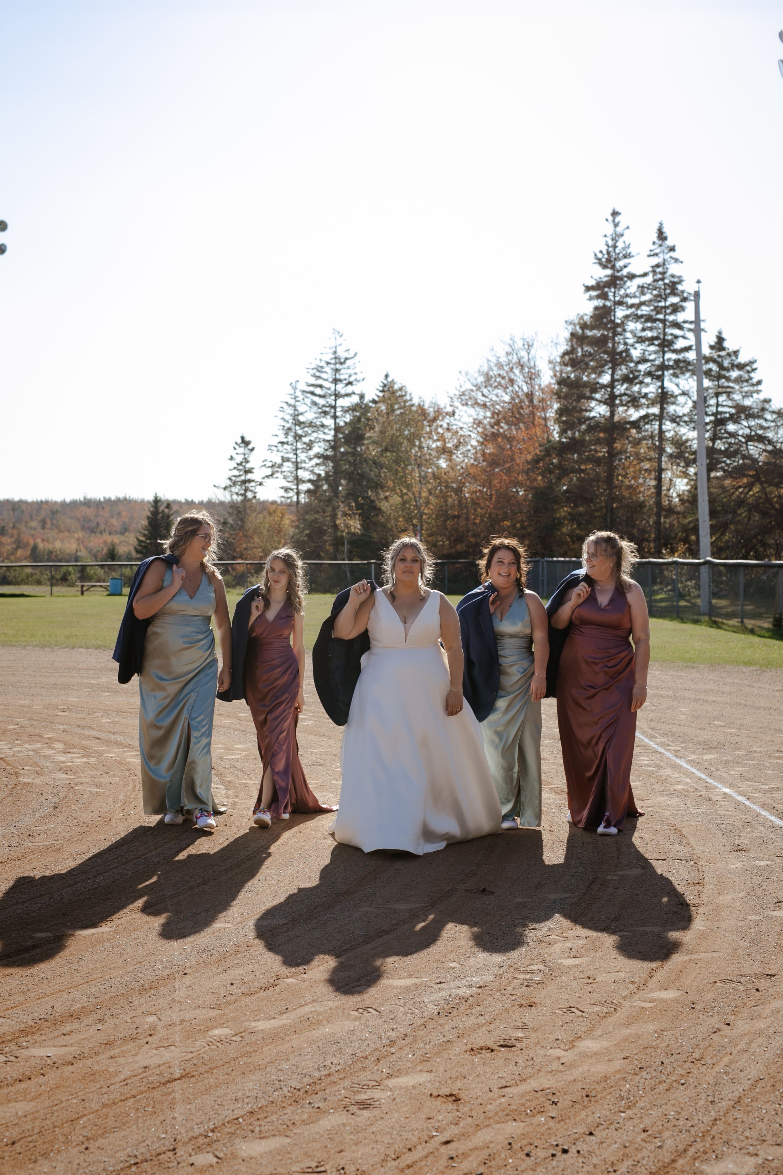 Bride and bridesmaids walking in baseball field. Photography by Samantha Bateman. Nova Scotia wedding photographer.