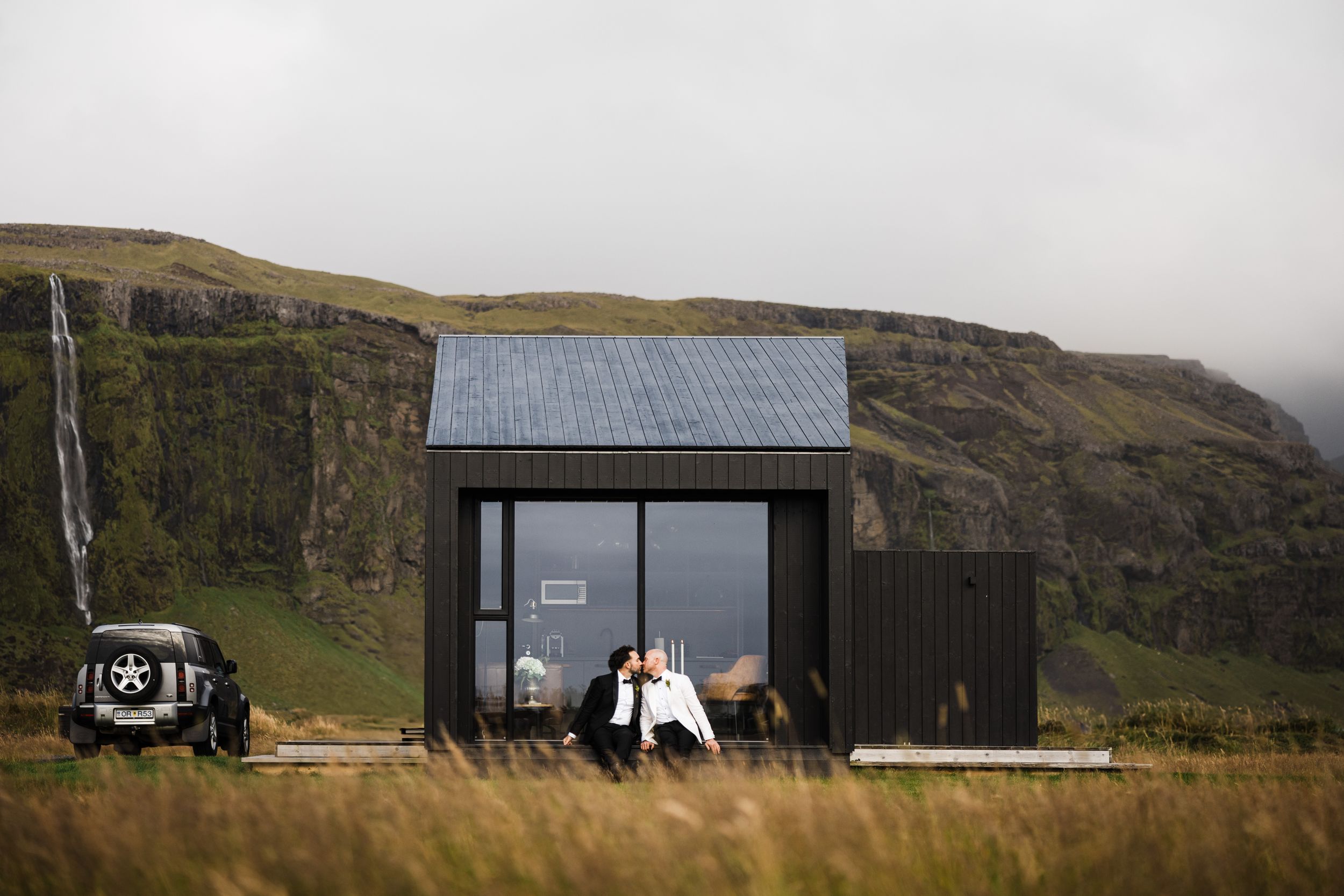 Modern black tiny house with solar panels sits against dramatic mountain backdrop in remote wilderness location.
