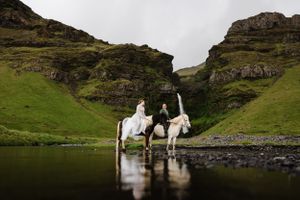 Two riders on white horses pose in front of a majestic waterfall in green mountains.
