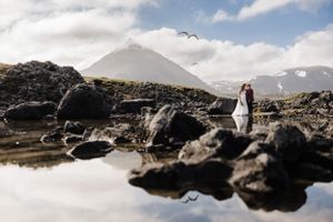 Wedding couple reflected in still water with mountain landscape and volcanic rocks.