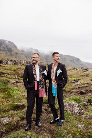 Two grooms in pinstripe suits with floral shirts pose on a rugged mountainside.
