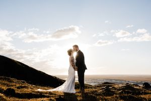 Two silhouetted figures in wedding attire embrace on a hilltop against a bright sunset sky over the ocean.