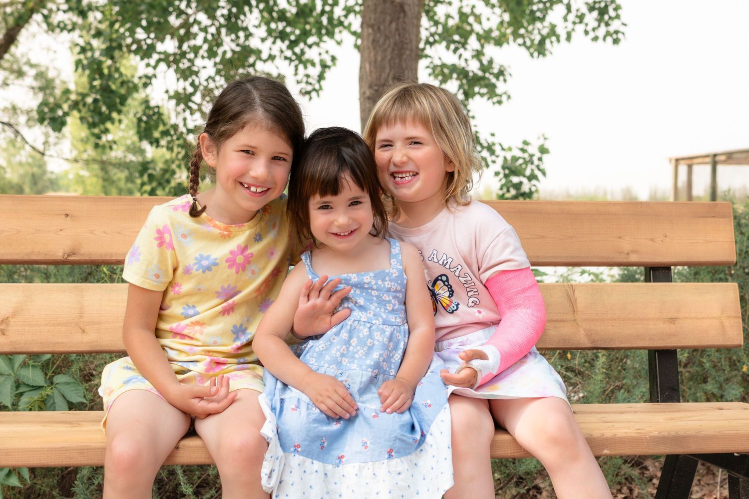 Three children sitting together on a wooden park bench outdoors on a sunny day.
