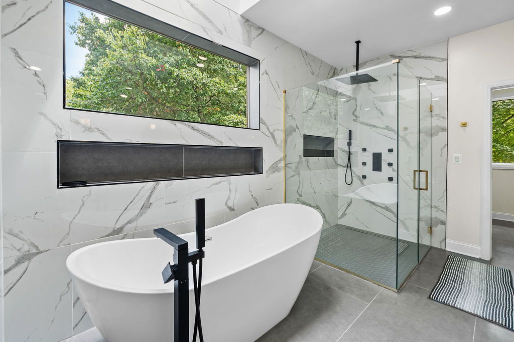 Modern luxury bathroom featuring a freestanding tub, glass shower enclosure, and marble walls with large window.