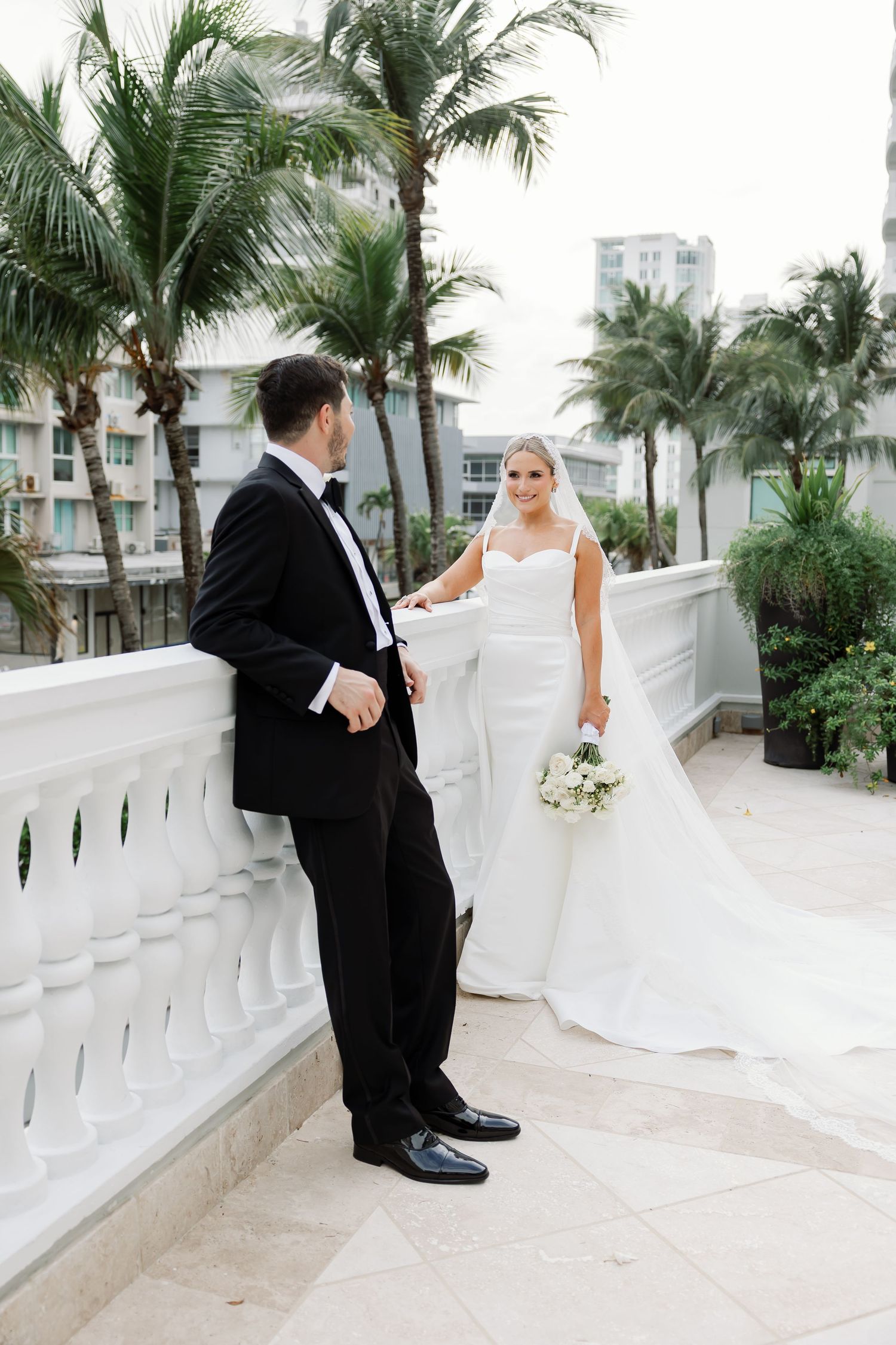 A bride and groom walk together on a grassy path with palm trees in the background.