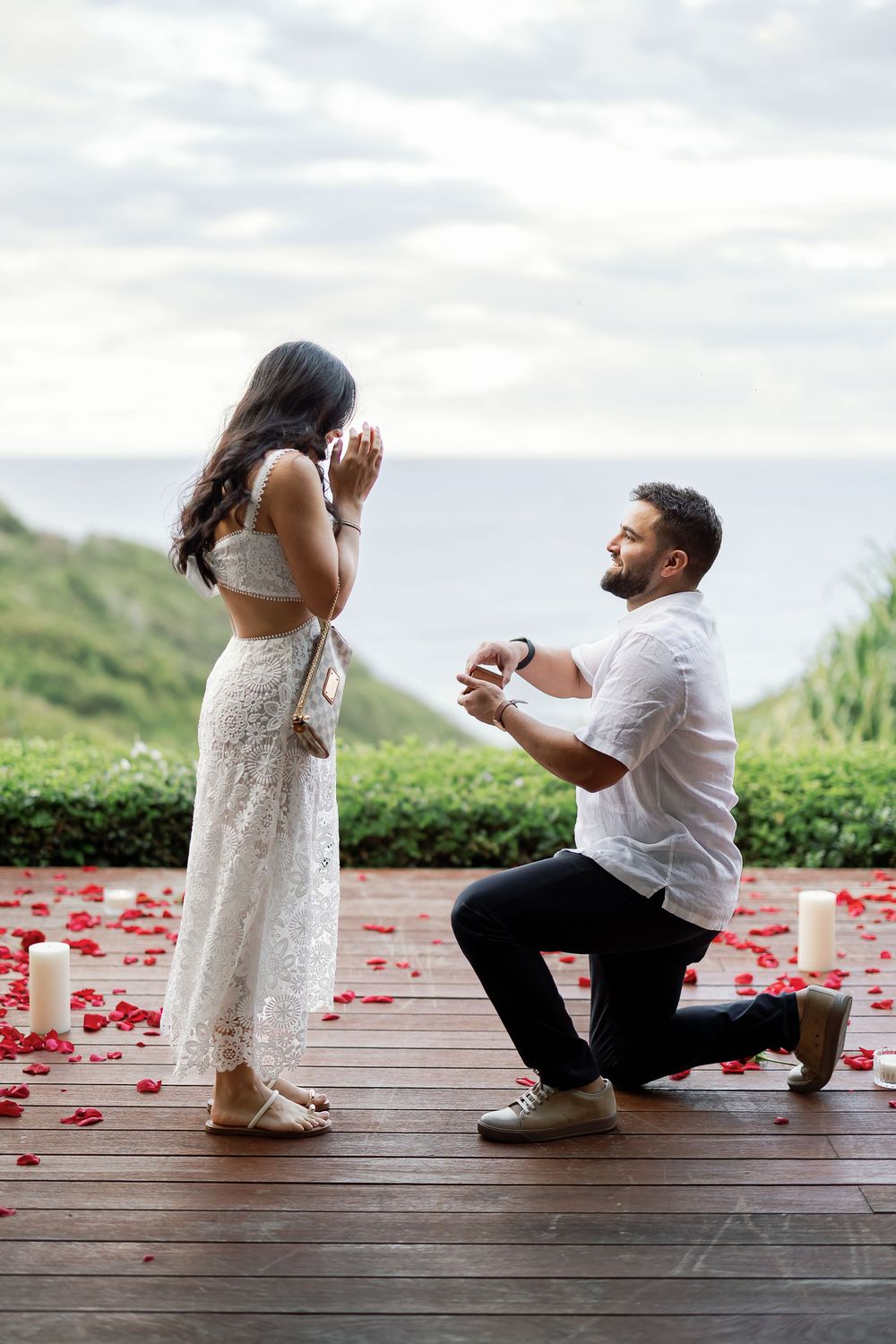A man kneeling on one knee and proposing to a woman on a wooden deck surrounded by rose petals.