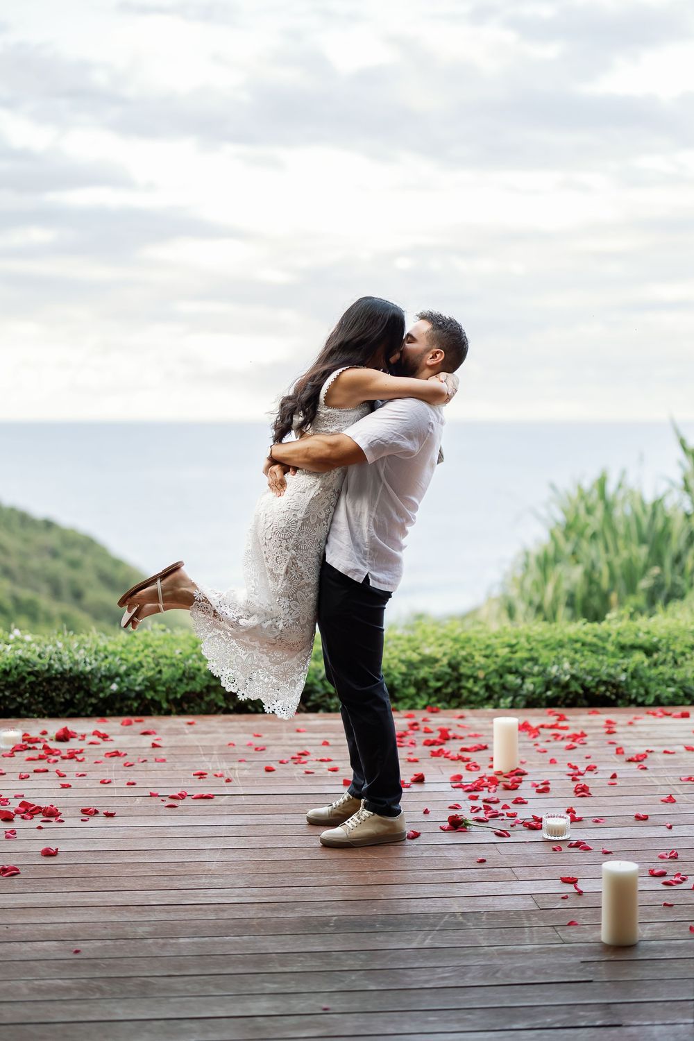 A couple embracing and kissing on a wooden deck with rose petals scattered around them.