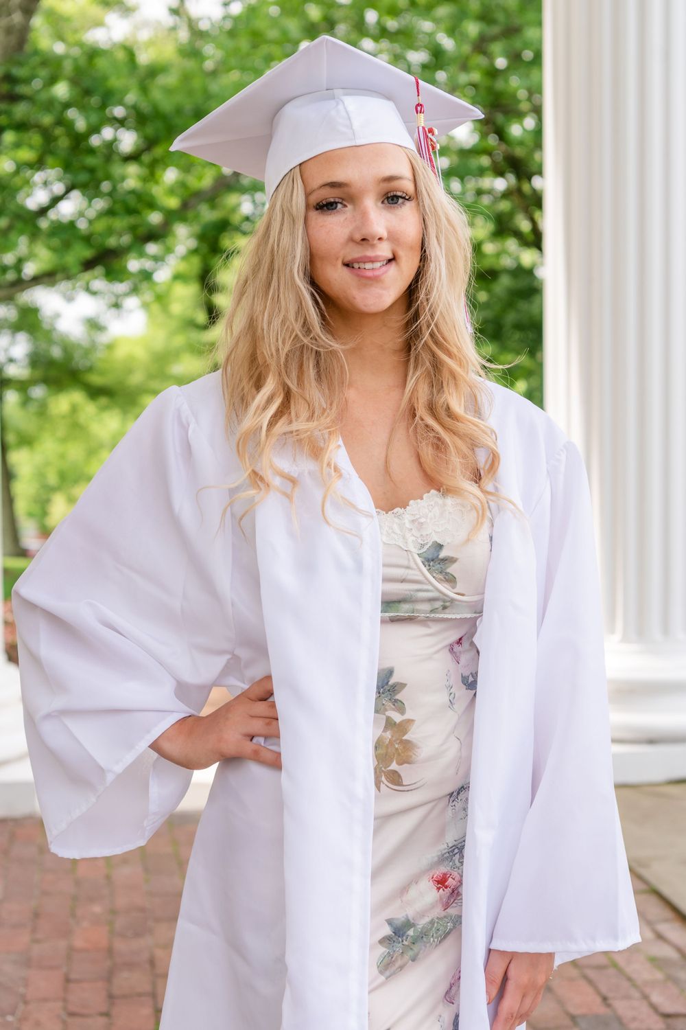 Graduate in white cap and gown wearing floral dress poses by classical column.