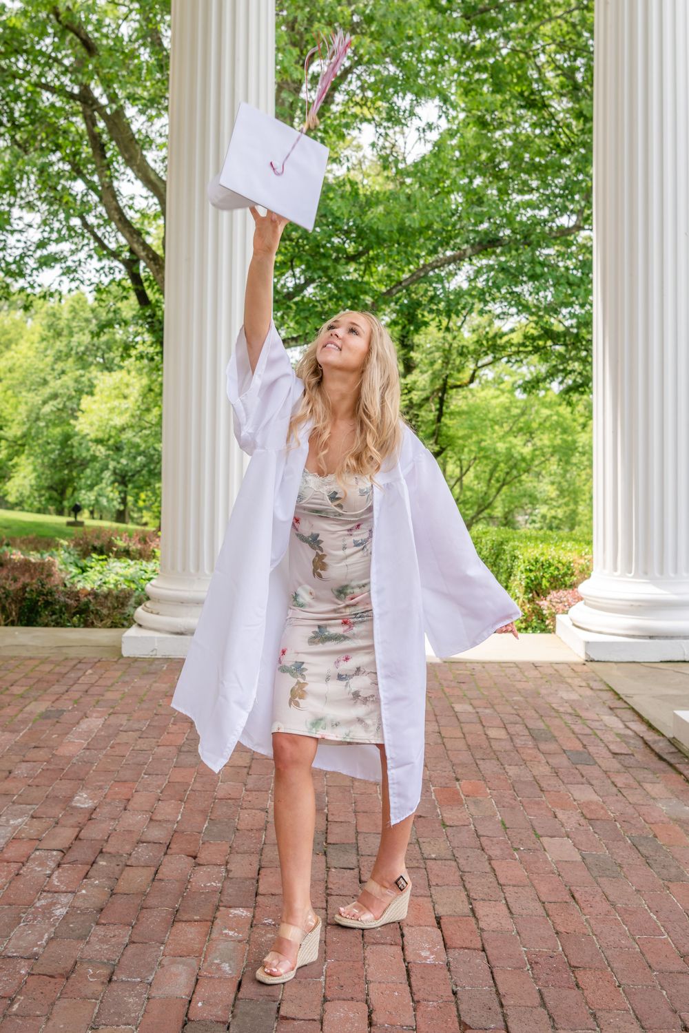 Graduate in white gown celebrating with diploma raised high between columns.