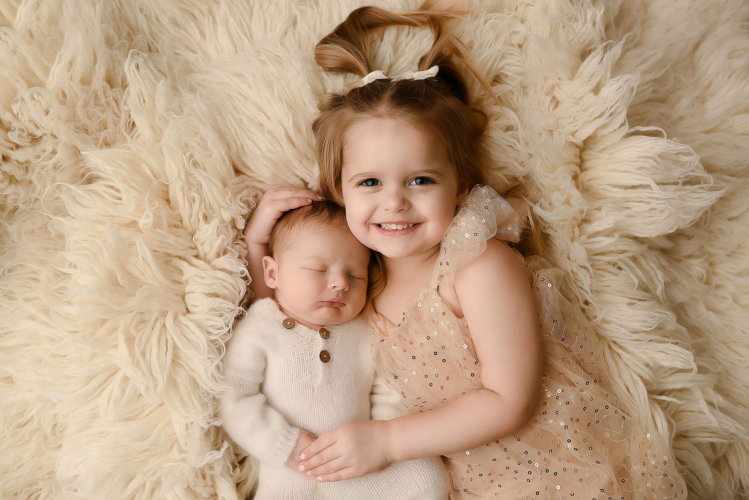 Smiling toddler girl in pink sparkle dress cuddling newborn sibling on a cream fluffy fur backdrop for sibling photos.