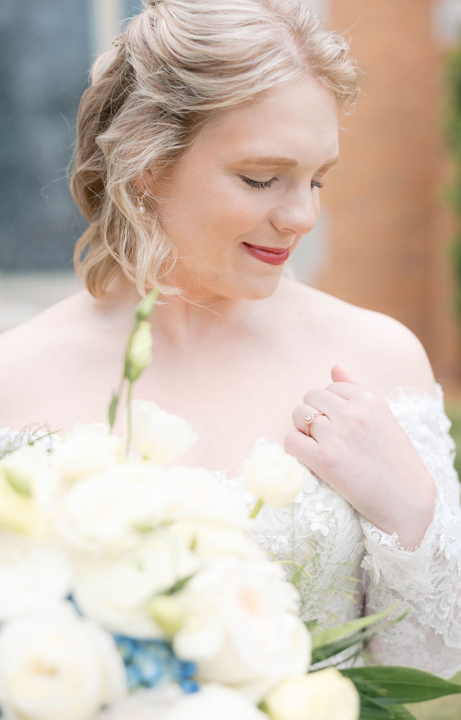 A bride in a lace gown holds white flowers, wearing a gold ring, with soft updo hair and red lips.