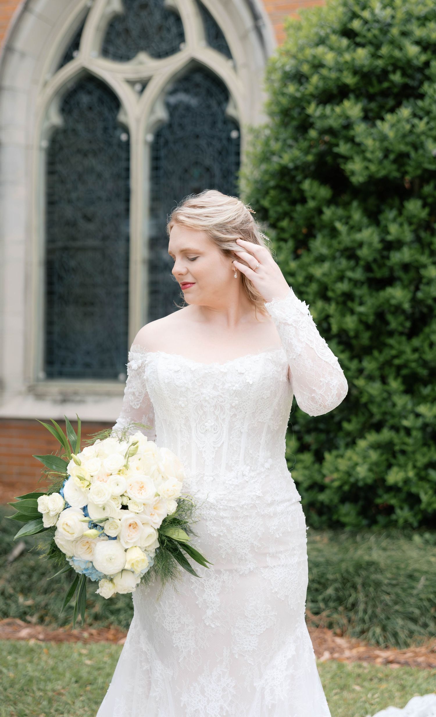 Bride in off-shoulder lace mermaid gown holds white rose bouquet outside a Gothic church.