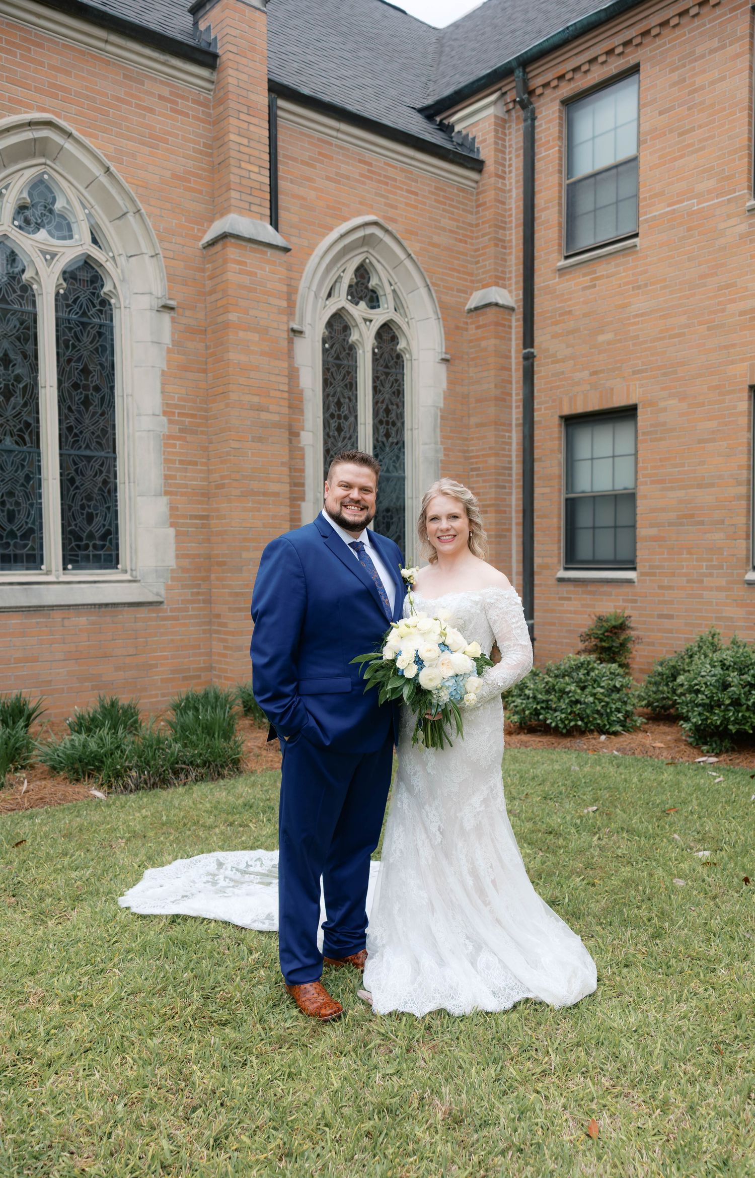 Bride and groom posing together outside a brick church, bride holding white bouquet in lace gown, groom in navy suit.