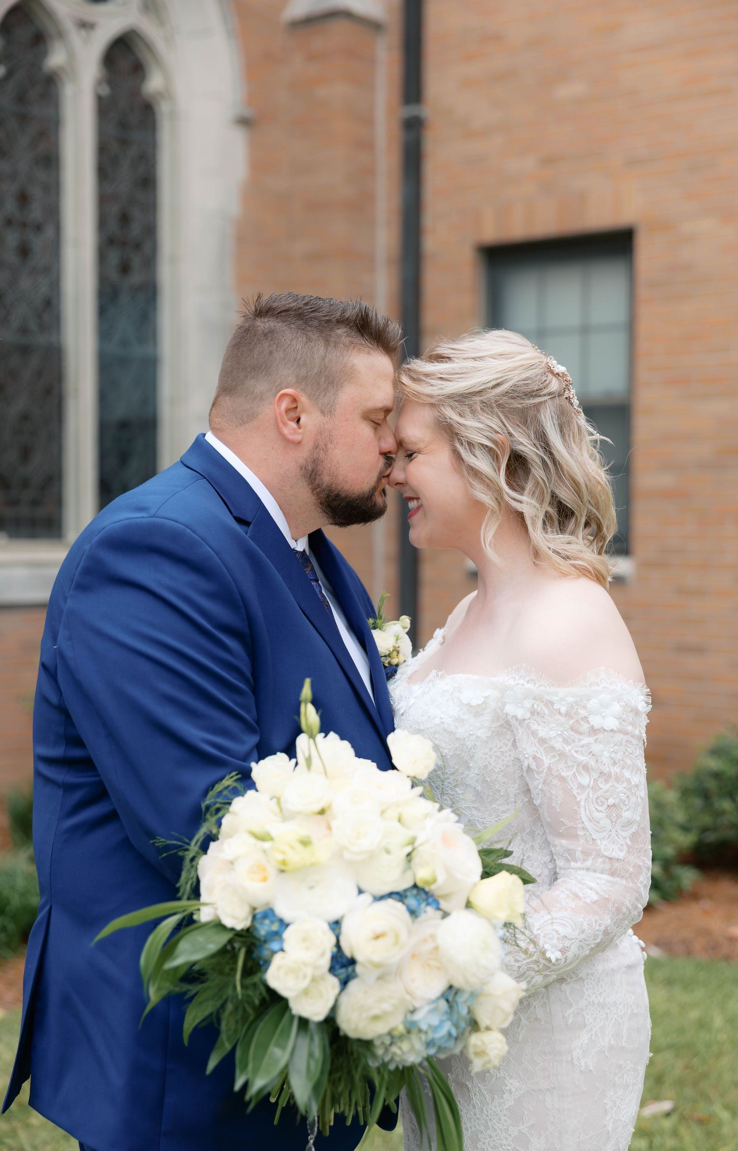 Bride and groom share a romantic forehead touch outside a church, bride holding white and blue floral bouquet.