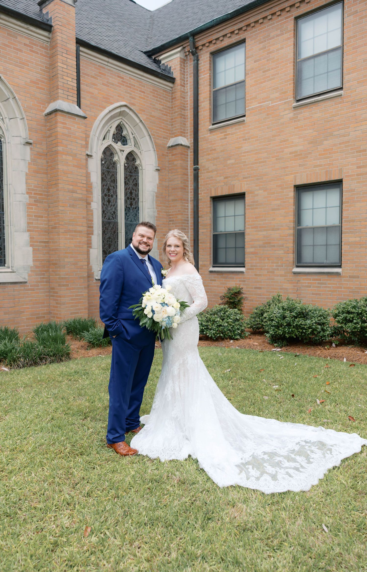 Bride and groom posing together outside a brick church, bride in lace gown with train, groom in navy suit.