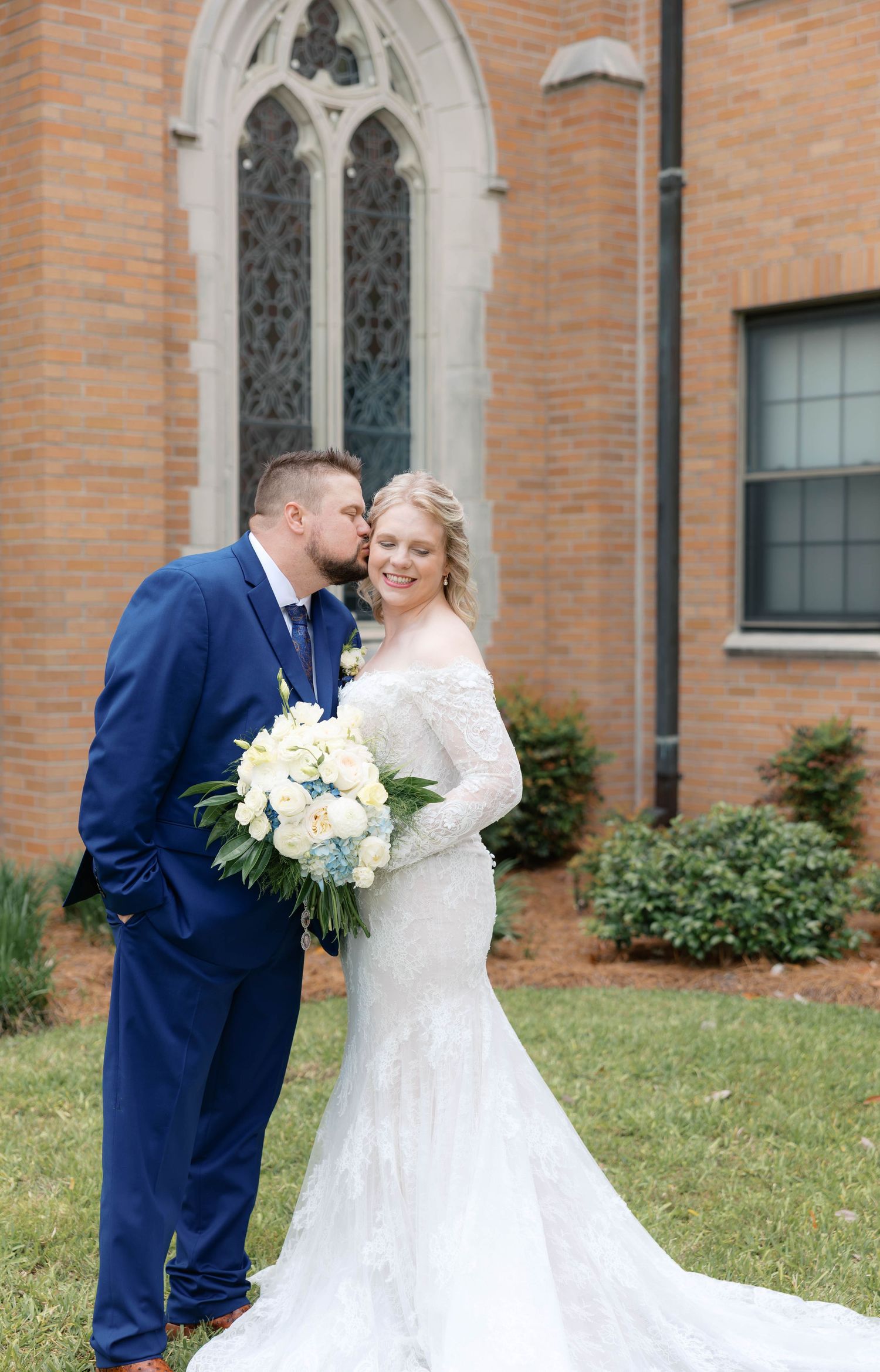 Bride and groom share a kiss outside a brick church, bride holding white and blue bouquet in lace mermaid gown.