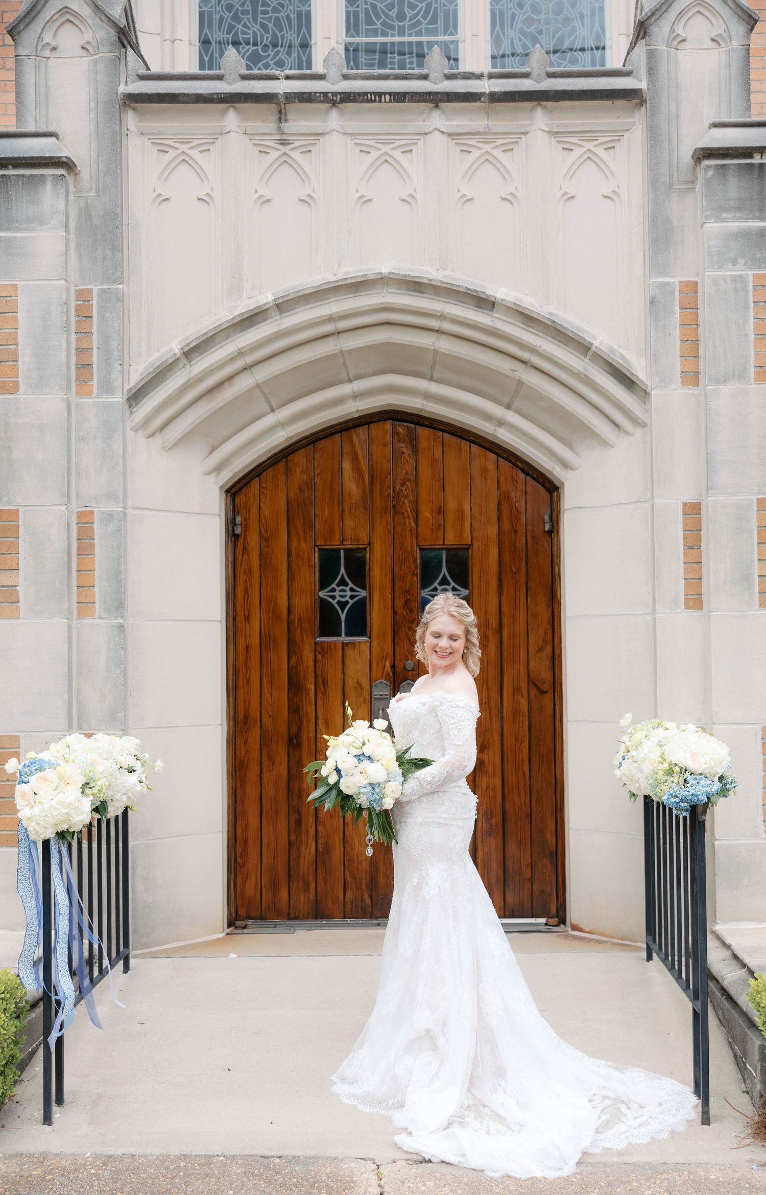 Bride in lace wedding gown holding white bouquet stands before ornate church wooden doors with floral arrangements.