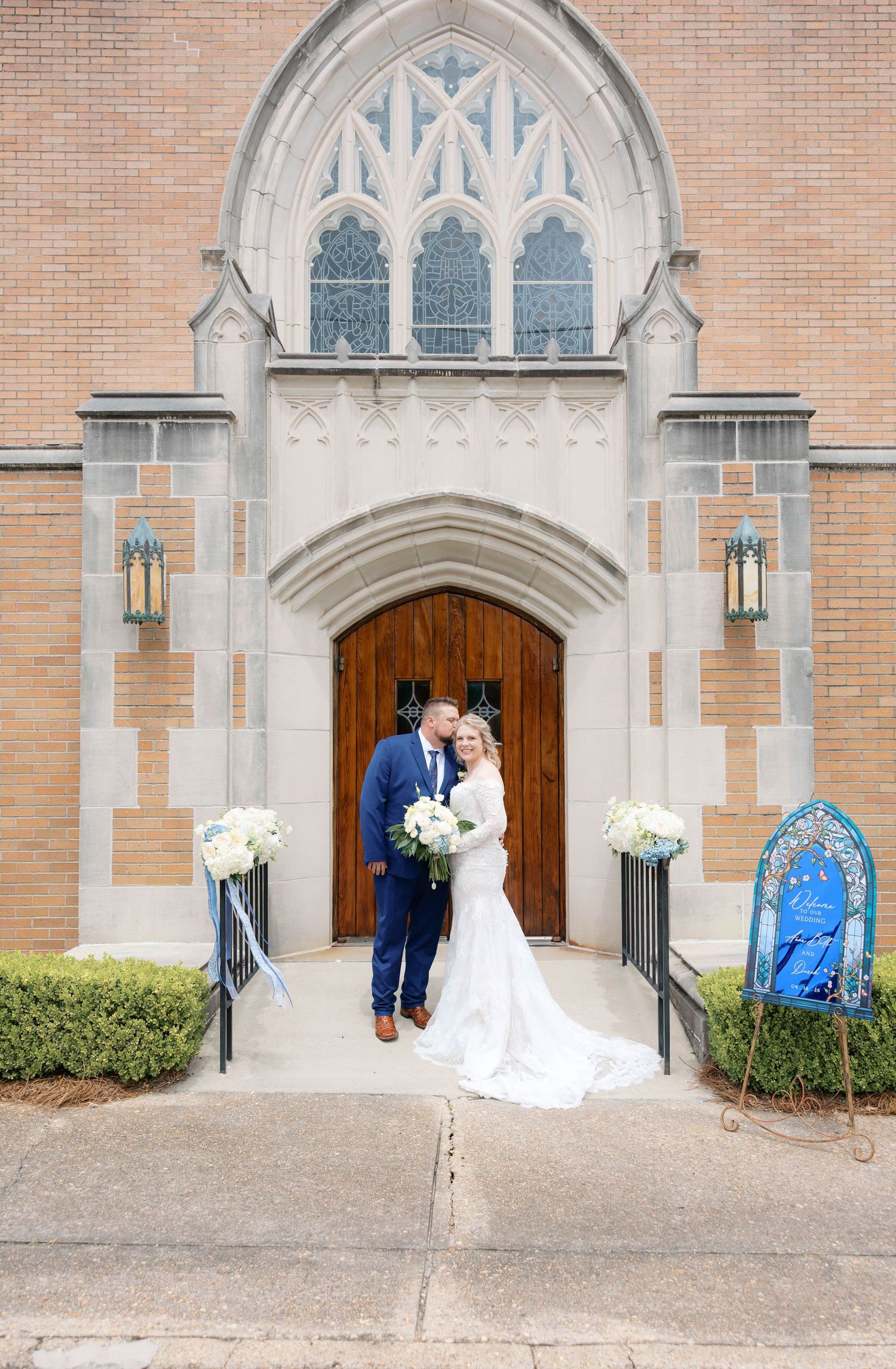 Bride and groom share romantic moment at Gothic church entrance with white floral arrangements and decorative signage.