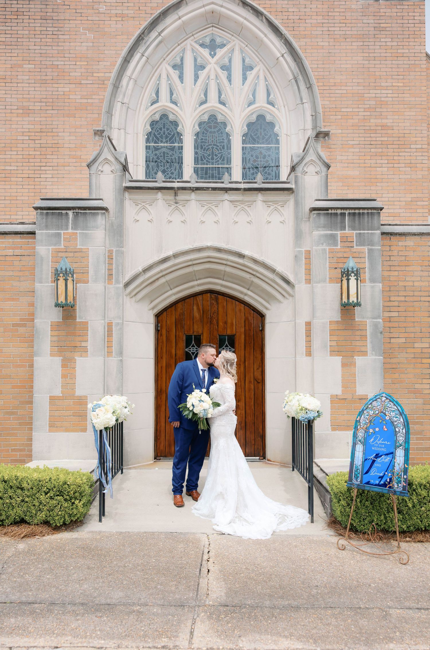Bride and groom share a romantic kiss at the entrance of a Gothic brick church with white floral decor.