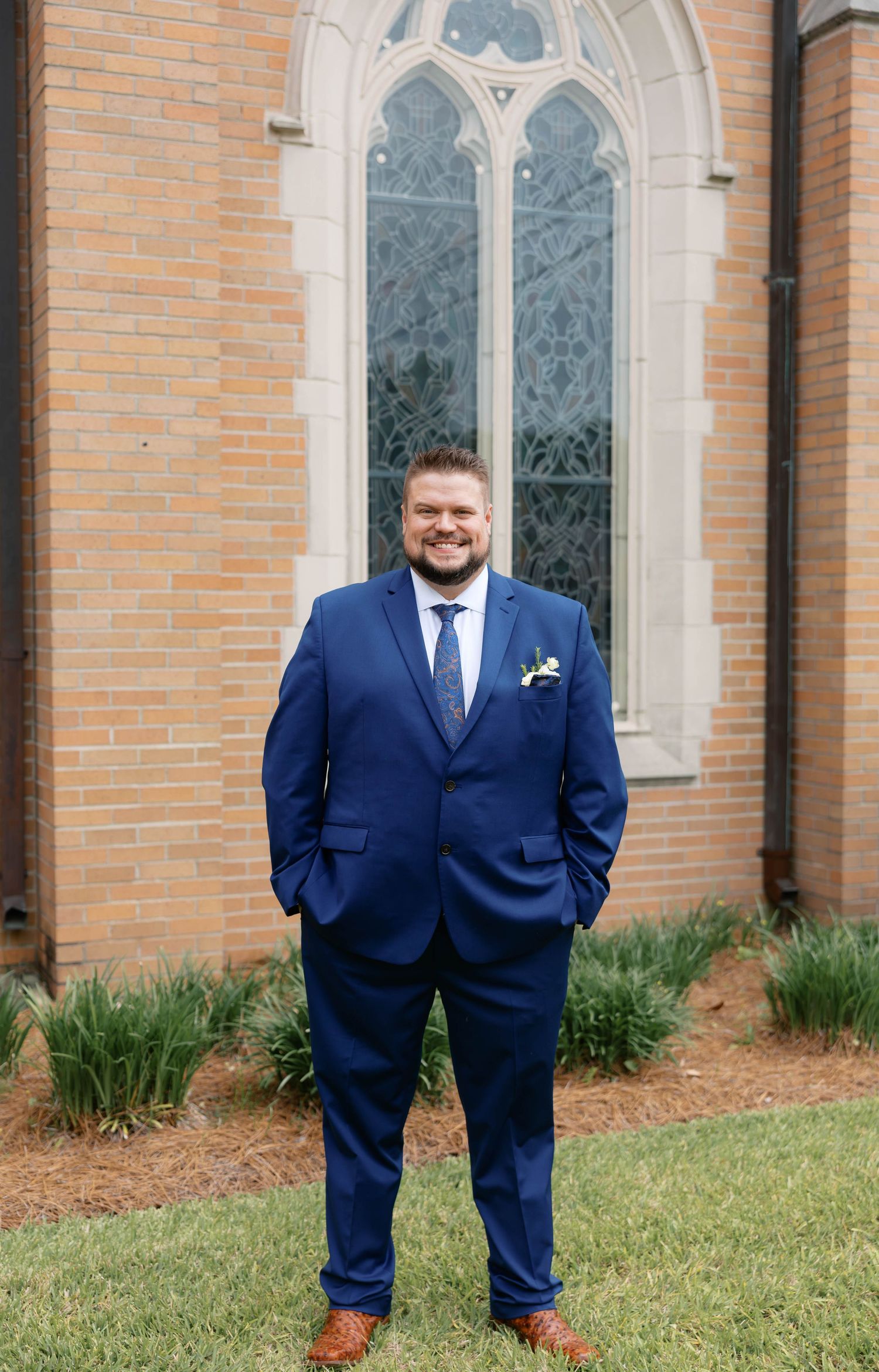 Smiling groom in navy blue suit with boutonniere stands outside brick church with Gothic stained glass window.