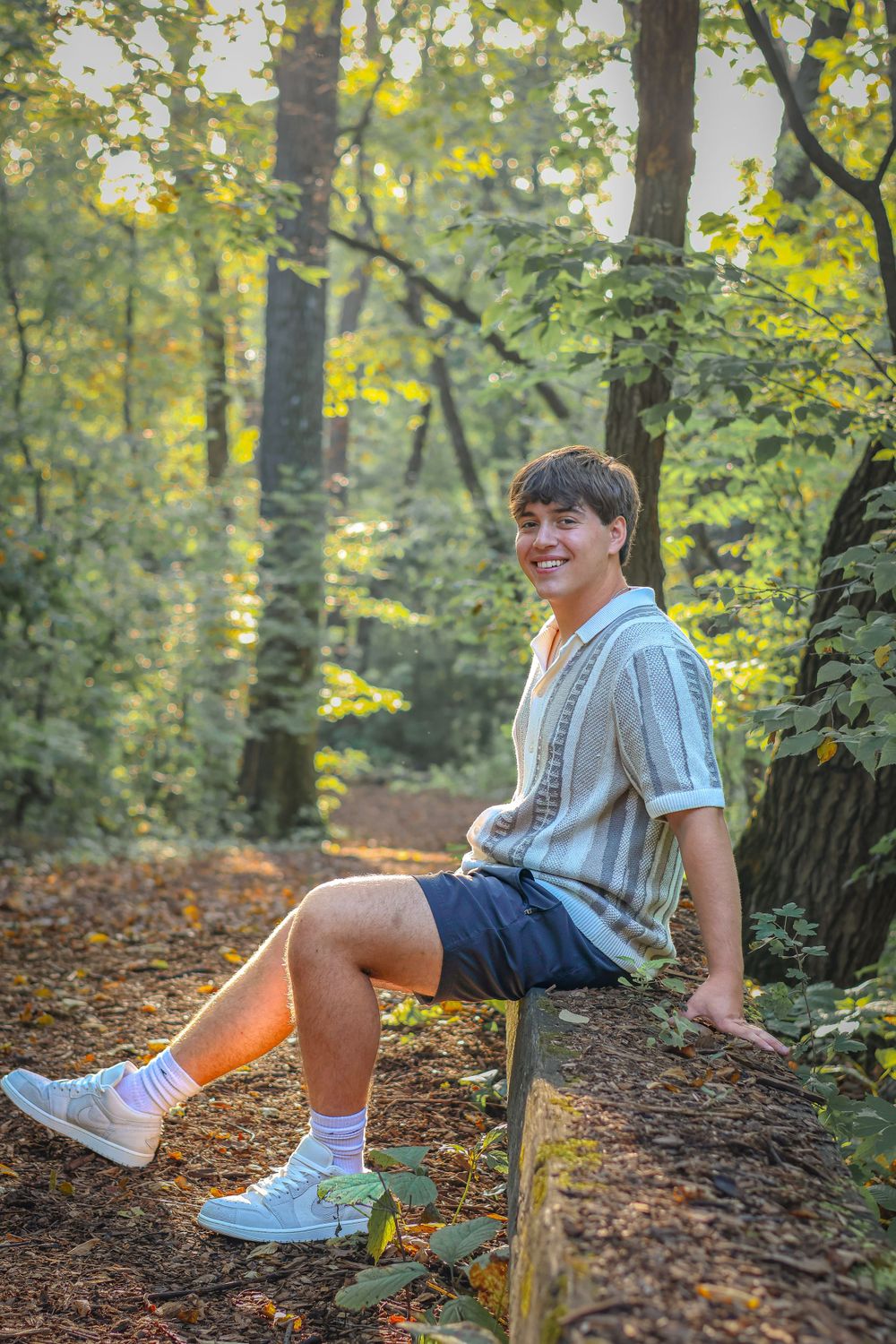 Person in light colored sweater perches on wooden railing along wooded trail.