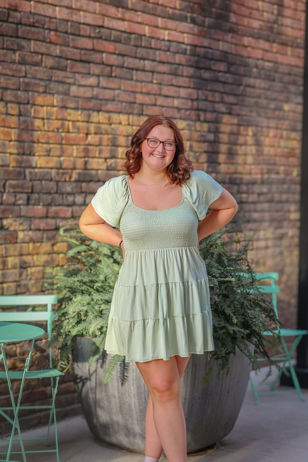 A summer portrait in a mint green dress beside a large planter with ferns and teal chairs.