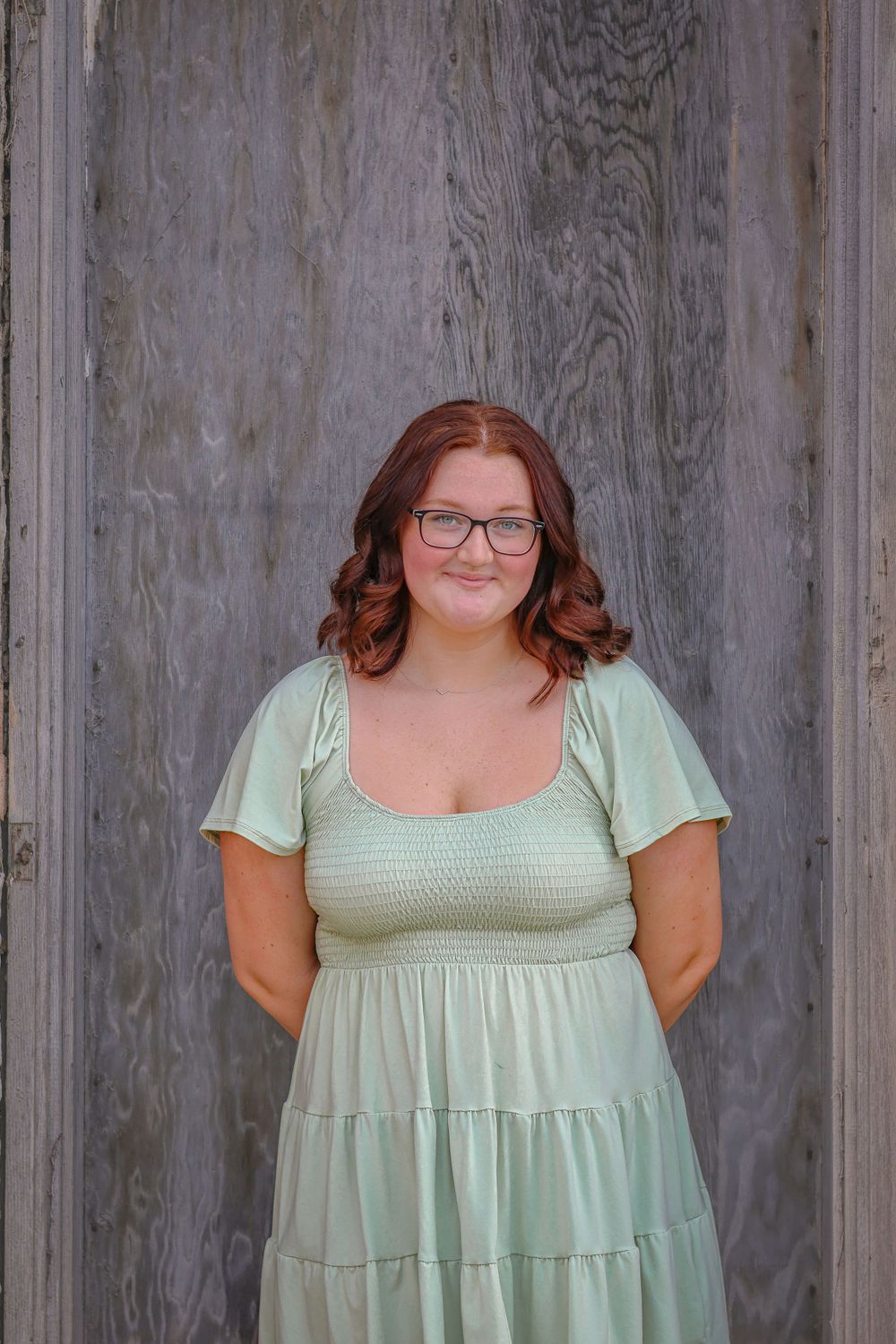 A portrait against a weathered wood wall wearing a mint green sundress and glasses.