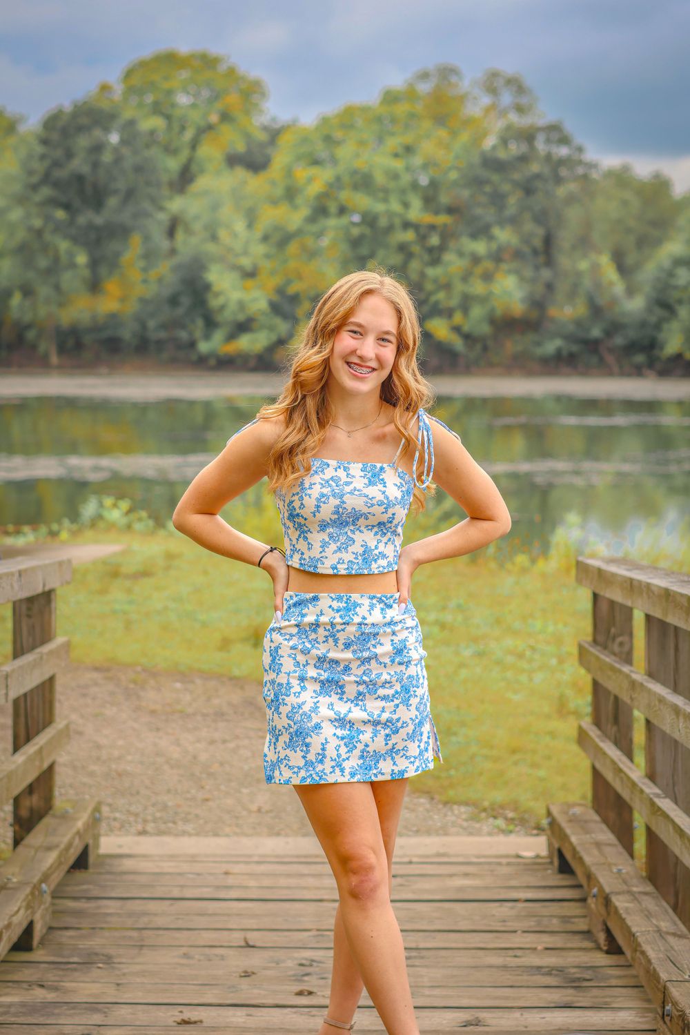 A portrait on a wooden bridge in a blue floral two-piece outfit with water and trees behind.