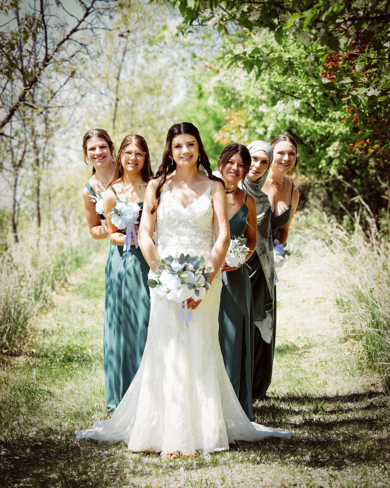 Wedding party in teal dresses poses with bride in white gown among trees and natural scenery.